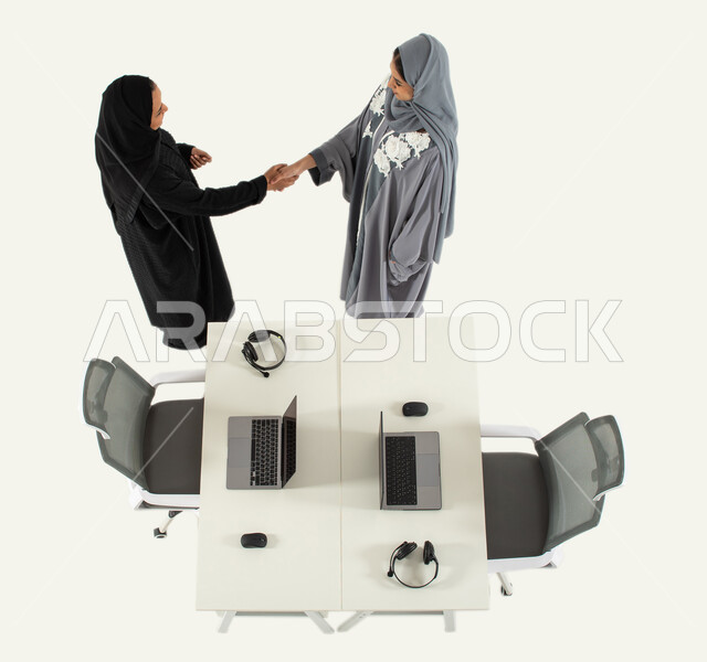 متابعة وتطوير العمل، صورة عامودية علوية لموظفتان عربيتان خليجيتان سعوديتان تتصافحان، استخدام الحاسوب المحمول، توقيع صفقات جديدة، اتفاقيات عمل ناجحة، العمل في الدعم الفني وخدمة العملاء، تقديم الاستشارات ومتابعة العمل، خلفية بيضاء