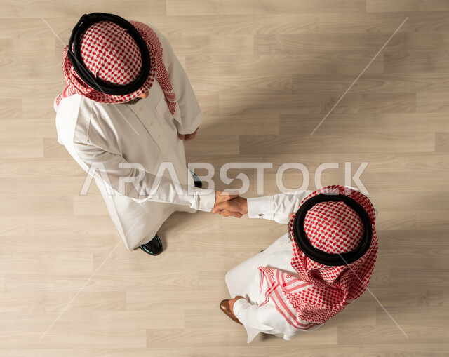 التصافح داخل الشركة ،عقد صفقات جديده ، اجتماع عمل , اتفاقيات عمل ناجحه ، صورة علوية عامودية لرجلان عربيان خليجيان سعوديان يرتديان الزي السعودي التقليدي , مهن ووظائف سعودية ، تحقيق اهداف الشركه