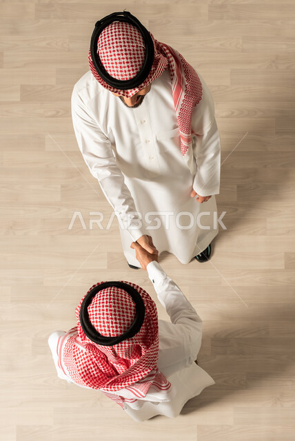 New deals, business meeting, successful business agreements, vertical overhead photo of two Saudi Arabian Gulf men wearing traditional Saudi dress, Saudi professions and jobs, shaking hands inside the company, achieving the company's goals