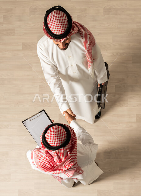 اجتماع عمل , اتفاقيات عمل ناجحه ، صورة علوية عامودية لرجلان عربيان خليجيان سعوديان يرتديان الزي السعودي التقليدي , مهن ووظائف سعودية ، التصافح داخل الشركة ،تحقيق اهداف الشركه ، عقد صفقات جديده