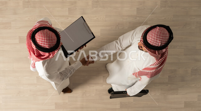 اتفاقيات عمل ناجحه ، صورة علوية عامودية لرجلان عربيان خليجيان سعوديان يرتديان الزي السعودي التقليدي , مهن ووظائف سعودية ، التصافح داخل الشركة ، اجتماع عمل , تحقيق اهداف الشركه ، عقد صفقات جديده