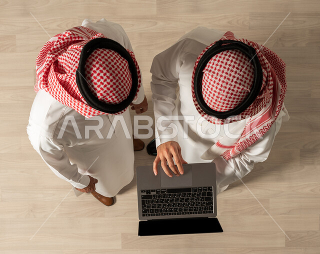 Joint group work, Saudi professions and jobs, a vertical overhead image of two Saudi Arabian Gulf men wearing traditional Saudi clothing, using a laptop, completing work tasks, achieving the company's goals, a business meeting