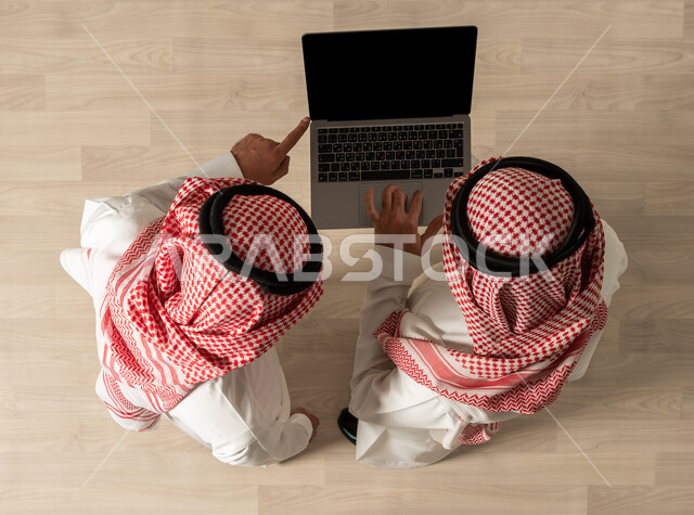 تحقيق اهداف الشركه ، اجتماع عمل , صورة علوية عامودية لرجلان عربيان خليجيان سعوديان يرتديان الزي السعودي التقليدي ، استخدام جهاز الحاسوب المحمول ، إنجاز مهام العمل ,مهن ووظائف سعودية