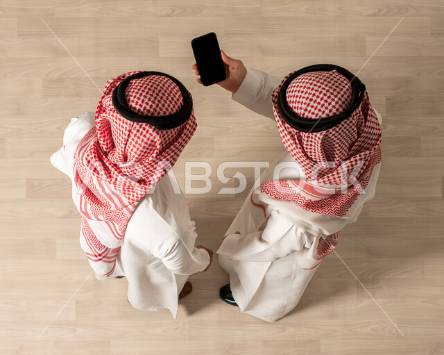 Using the mobile phone to complete work tasks, a vertical overhead image of two Saudi Arab Gulf men wearing traditional Saudi dress, completing work tasks, co-workers, Saudi professions and jobs, a business meeting, achieving the company's goals