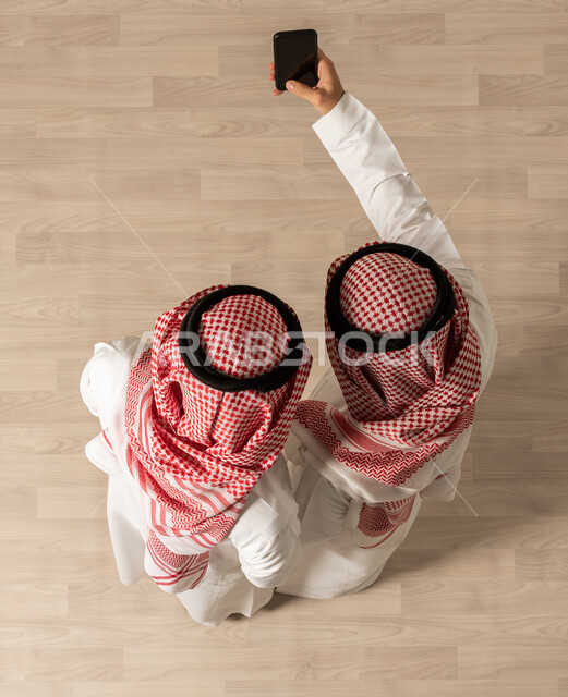 التقاط صور سلفي عبر الهاتف المحمول ، صورة علوية عامودية لرجلان عربيان خليجيان سعوديان يرتديان الزي السعودي التقليدي ، إنجاز مهام العمل  ، مهن ووظائف سعودية ، اجتماع عمل , تحقيق اهداف الشركه