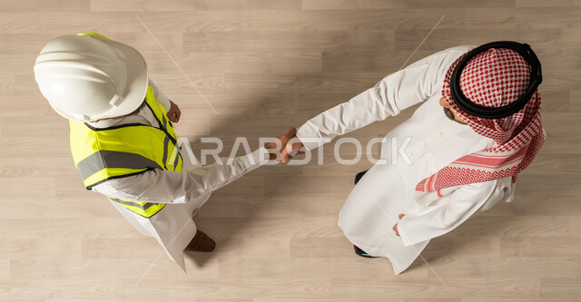 عقد صفقات جديده, اتفاقيات عمل ناجحه ، التحية والسلام ، صورة علوية عامودية لرجلان عربيان خليجيان سعوديان يرتديان الزي السعودي التقليدي ، اجتماع عمل مع مهندس معماري يرتدي البدلة وخوذة السلامة ، التصافح في مقر الشركة ، مهن ووظائف سعودية