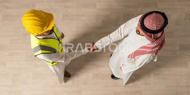 Shaking hands at the company's headquarters, a vertical overhead image of two Saudi Arabian Gulf men wearing the traditional Saudi dress, a business meeting with an architect wearing a suit and a safety helmet, salutations and peace, Saudi professions and