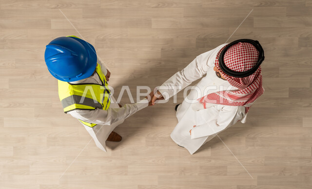 العمل مع الأصدقاء ،صورة علوية عامودية لرجلان عربيان خليجيان سعوديان يرتديان الزي السعودي التقليدي ، اجتماع عمل مع مهندس معماري يرتدي البدلة وخوذة السلامة  , مهن ووظائف سعودية ، عقد صفقات جديده, اتفاقيات عمل ناجحه