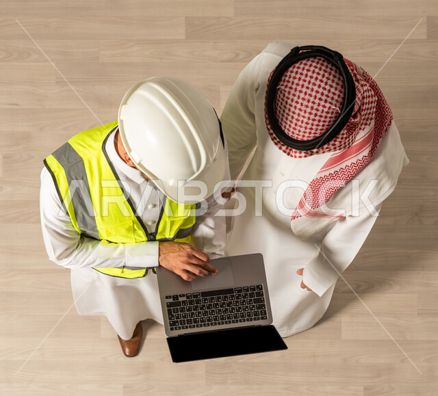 إنجاز مهام العمل , نقاشات عمل ، صورة علوية عامودية لرجلان عربيان خليجيان سعوديان يرتديان الزي السعودي التقليدي , استخدام جهاز الحاسوب المحمول ,  اجتماع عمل مع مهندس معماري يرتدي البدلة وخوذة السلامة ،  مهن ووظائف سعودية