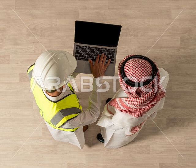  اجتماع عمل مع مهندس معماري يرتدي البدلة وخوذة السلامة ، صورة علوية عامودية لرجلان عربيان خليجيان سعوديان يرتديان الزي السعودي التقليدي , استخدام جهاز الحاسوب المحمول , إنجاز مهام العمل , نقاشات عمل