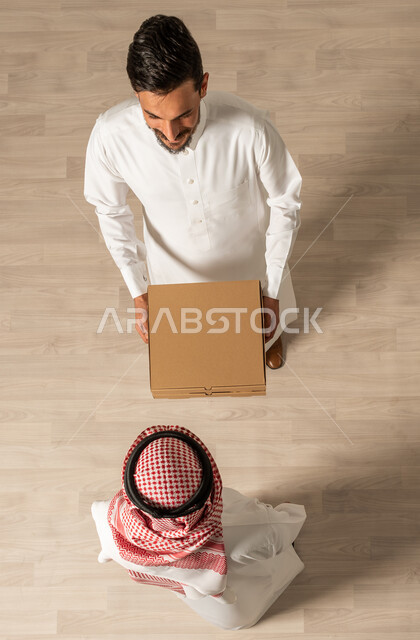 طلبات اونلاين, التسوق الذكي ، صورة علوية عامودية لشابان عربيان خليجيان سعوديان يرتديان الزي السعودي التقليدي ، عملية شحن وتوصيل موثوقه, خدمة التوصيل السريع ،  التسوق الالكتروني 