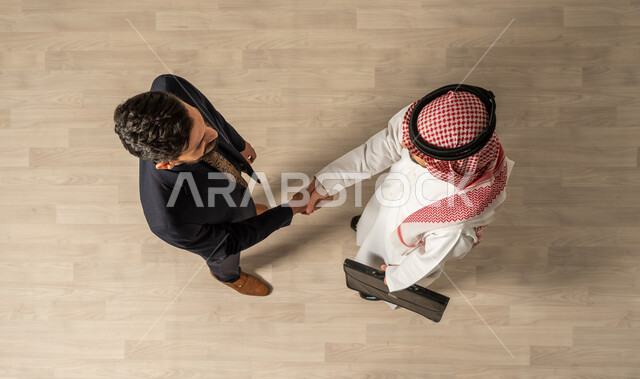 التحية والسلام ، صوره علويه عاموديه لرجل عربي سعودي خليجي يرتدي الزي السعودي التقليدي ورجل عربي يرتدي البدله الرسميه ، التصافح في مقر الشركة ، لقاء الأصدقاء ، مهن ووظائف السعودية ، صفقة عمل ناجحة