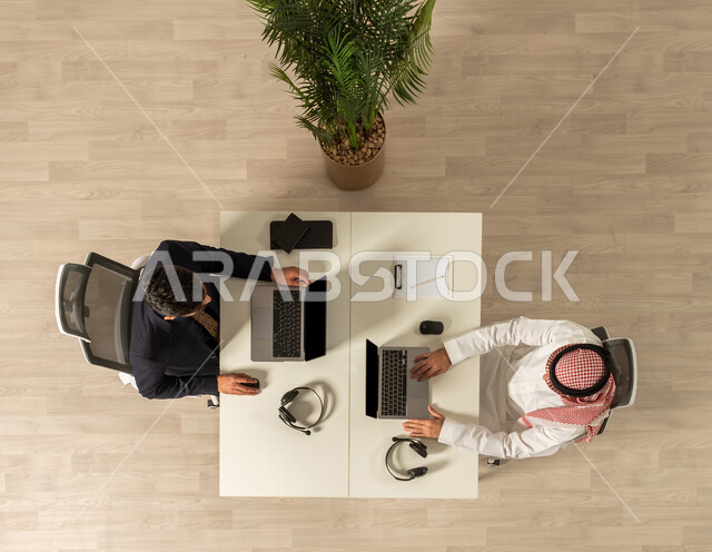 مهن ووظائف سعوديه ، صوره علويه عاموديه لرجل عربي خليجي سعودي يرتدي الزي التقليدي ورجل يرتدي البدلة الرسمية في مقر الشركة ، استخدام جهاز الحاسوب المحمول، انجاز مهام العمل، اعمال مكتبيه