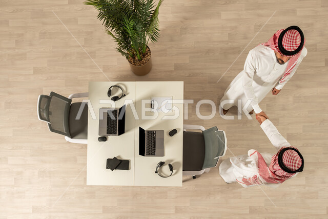 عمل جماعي مشترك ، صورة علوية عامودية لرجلان عربيان خليجيان سعوديان يرتديان الزي السعودي التقليدي ، التصافح داخل الشركة ،  اتفاق عمل بين رجال الاعمال ، صفقة ناجحة ، تعاون تجاري 