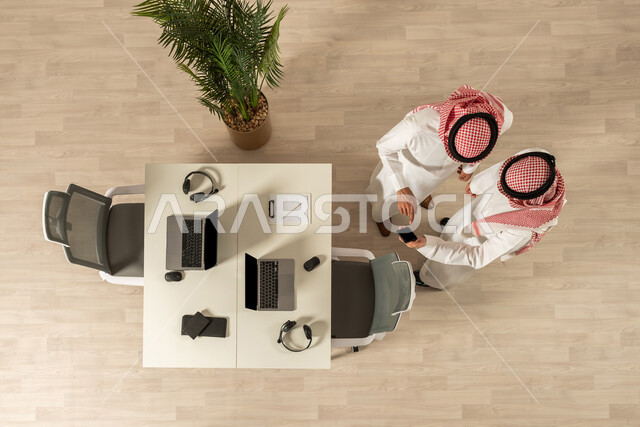 استخدام الهاتف المحمول  ،صورة علوية عامودية لرجلان عربيان خليجيان سعوديان يرتديان الزي السعودي التقليدي ، نقاشات وحوارات ، اجتماع الزملاء في مقر العمل ، تحقيق أهداف الشركة ، عمل جماعي مشترك