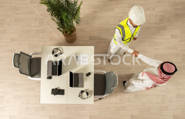اتفاق عمل بين رجال الاعمال ،صورة علوية عامودية لرجلان عربيان خليجيان سعوديان يرتديان الزي السعودي التقليدي ، التصافح داخل الشركة ،  اجتماع عمل مع مهندس معماري يرتدي البدلة وخوذة السلامة ، صفقة ناجحة ، تعاون تجاري ، عمل جماعي مشترك