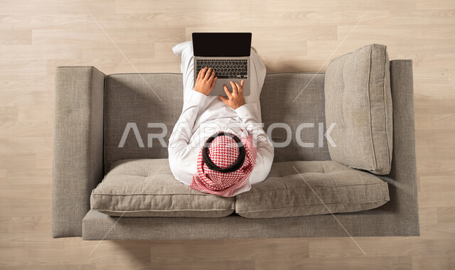 صورة من الاعلى لرجل عربي خليجي سعودي يجلس على الاريكة ويستخدم جهاز الحاسوب المحمول، غرفة المعيشة داخل المنزل الجديد، العمل عن بعد وتصفح الانترنت، انجاز مهام العمل، احتراف وظائف الحاسوب وتكنولوجيا المعلومات