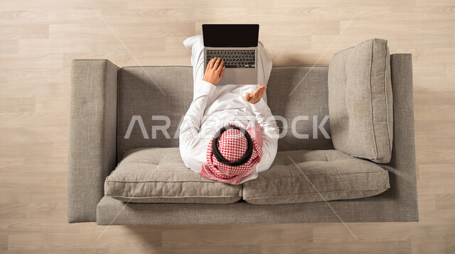 صورة من الاعلى لرجل عربي خليجي سعودي يجلس على الاريكة ويقوم باجراءة مكالمة فيديو من خلال الحاسوب المحمول، اجتماع عمل عن بعد، القاء التحية من خلال شاشة اللابتوب، غرفة المعيشة داخل المنزل الجديد، انجاز مهام العمل، احتراف وظائف الحاسوب