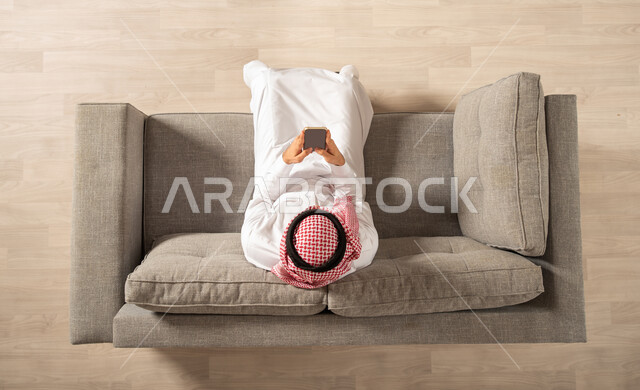 صورة من الاعلى لرجل عربي خليجي سعودي يجلس على الاريكة في المنزل الجديد ويستخدم الهاتف المحمول، امتلاك بيت جديد، تصفح الانترنت، استخدام وسائل التواصل الاجتماعي، بث فيديو مباشر، اجهزة تقنية حديثة