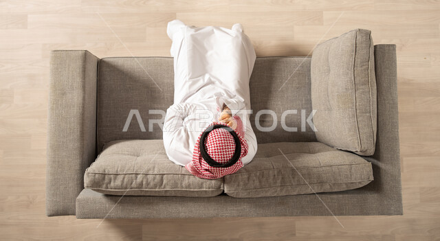 صورة علوية عامودية لرجل عربي سعودي خليجي  في غرفة المعيشة داخل المنزل الجديد ، امتلاك سكن ،  الجلوس على الاريكة ,ايماءات تدل على السعادة والرضى ،   منازل و عقارات  .