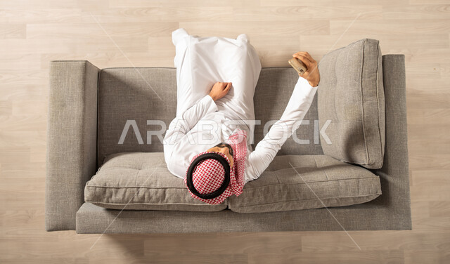 صورة من الاعلى لرجل عربي خليجي سعودي يجلس على الاريكة في المنزل الجديد ويستخدم الهاتف المحمول، التقاط صور تذكارية، بث فيديو مباشر، اجراء مكالمة فيديو، اجهزة تقنية حديثة، امتلاك بيت جديد