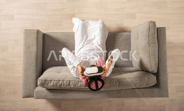 رجل عربي خليجي سعودي يجلس على الاريكة ويرتدي نظارة الواقع الإفتراضي ثلاثية الأبعاد، التفاعل مع الواقع الافتراضي، المتعة والتسلية بمشاهدة الأفلام ومحاكاة العالم الإفتراضي، الاستقرار في المنزل الجديد