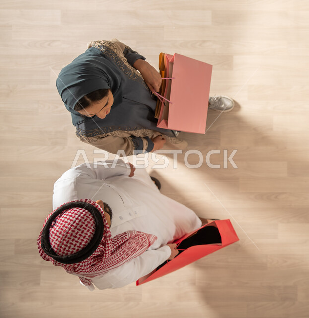 A picture from above of a Saudi Arab Gulf couple carrying shopping bags, returning from the market, buying the necessary items, benefiting from discounts and sales, Black and White Friday, sales and sales, the pleasure of buying and shopping in Saudi Arab