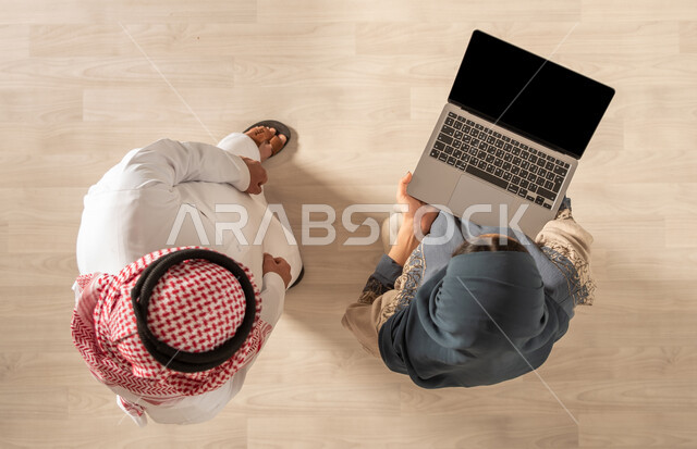 صورة من الاعلى لرجل وامرأة عربيان خليجيان سعوديان يرتديان الزي السعودي التقليدي ويستخدمان الحاسوب المحمول، متابعة ومناقشة الاعمال، جلسة عمل داخل مقر الشركة، بيئة العمل السعودي، فريق عمل سعودي، زملاء عمل
