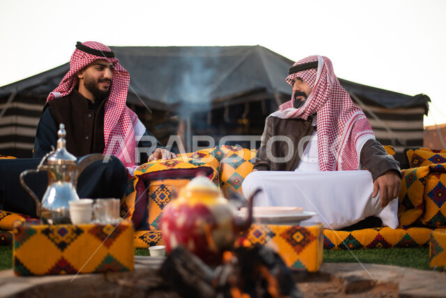 قضاء أوقات ممتعة مع الأصحاب، أجواء تخييم شبابية ليلية أمام الحطب، رجلان عربيان خليجيان سعوديان يرتديان الثوب التقليدي والشماغ مع ملابس الشتاء الدافئة يجلسون أمام النيران المشتعلة في كشتة برية، جلسة بر عربية شعبية، حسن الضيافة والاستقبال عند العرب