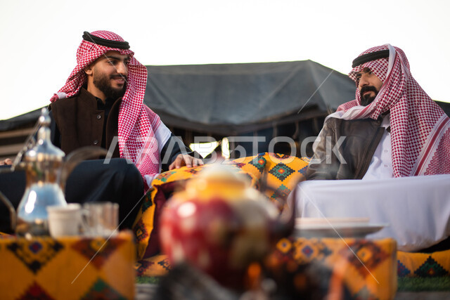 كشتة بر شبابية، الأجواء الهادئة الشتوية في المخيمات الشعبيه في صحاري السعودية، علاقات إجتماعية بين الأصحاب، جلسة وناسة وتعارف، دردشات وحوارات بين الاصدقاء، رجلان عربيان خليجيان سعوديان يرتديان الثوب التقليدي والشماغ مع ملابس الشتاء الدافئة يقضون أوقات ممتعة