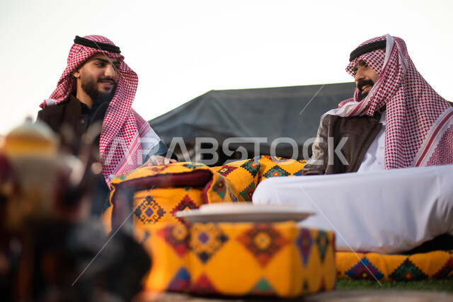 الأجواء الهادئة الشتوية في المخيمات الشعبيه في صحاري السعودية، علاقات إجتماعية بين الأصحاب، جلسة وناسة وتعارف، دردشات وحوارات بين الاصدقاء، رجلان عربيان خليجيان سعوديان يرتديان الثوب التقليدي والشماغ مع ملابس الشتاء الدافئة يقضون أوقات ممتعة،  كشتة بر شبابية