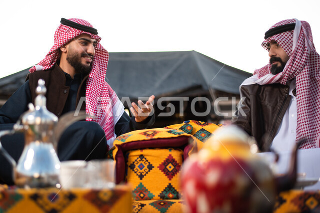 علاقات إجتماعية بين الأصحاب، جلسة وناسة وتعارف، دردشات وحوارات بين الاصدقاء، رجلان عربيان خليجيان سعوديان يرتديان الثوب التقليدي والشماغ مع ملابس الشتاء الدافئة يقضون أوقات ممتعة،  كشتة بر شبابية، الأجواء الهادئة الشتوية في المخيمات الشعبيه في صحاري السعودية
