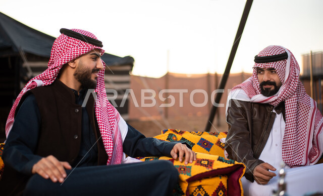 جلسة وناسة وتعارف، دردشات وحوارات بين الاصدقاء، رجلان عربيان خليجيان سعوديان يرتديان الثوب التقليدي والشماغ مع ملابس الشتاء الدافئة يقضون أوقات ممتعة،  كشتة بر شبابية، الأجواء الهادئة الشتوية في المخيمات الشعبيه في صحاري السعودية، علاقات إجتماعية بين الأصحاب