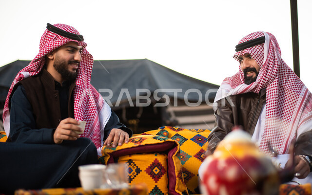كشتة بر شبابية مسائية، اجواء شتوية ليلية، مفهوم حسن الضيافه والكرم، شابان عربيان خليجيان سعوديان يرتديان الثوب التقليدي والشماغ مع ملابس الشتاء في رحلة تخييم برية في صحراء السعودية، تقديم القهوة العربية الساخنة للضيوف، حوارات ونقاشات بين الاصدقاء