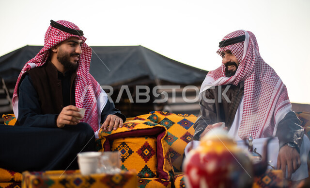 اجواء شتوية ليلية، مفهوم حسن الضيافه والكرم، شابان عربيان خليجيان سعوديان يرتديان الثوب التقليدي والشماغ مع ملابس الشتاء في رحلة تخييم برية في صحراء السعودية، تقديم القهوة العربية الساخنة للضيوف، حوارات ونقاشات بين الاصدقاء، كشتة بر شبابية مسائية