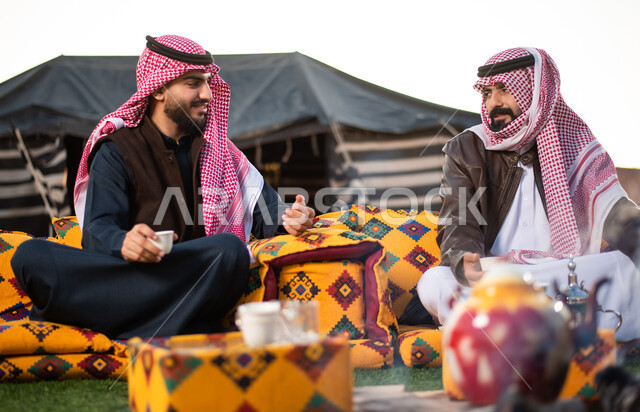 مفهوم حسن الضيافه والكرم، شابان عربيان خليجيان سعوديان يرتديان الثوب التقليدي والشماغ مع ملابس الشتاء في رحلة تخييم برية في صحراء السعودية، تقديم القهوة العربية الساخنة للضيوف، حوارات ونقاشات بين الاصدقاء، كشتة بر شبابية مسائية، اجواء شتوية ليلية