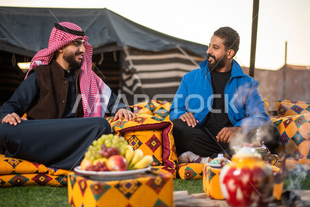 Good hospitality and reception, a youth camping atmosphere at night in front of a firewood flame, Saudi Gulf Arab friends sitting in front of the burning fires in a wild kashta, a popular Arab bar session, tourist camps in the deserts of Saudi Arabia, enjoying spending time with friends, wearing warm winter clothes.