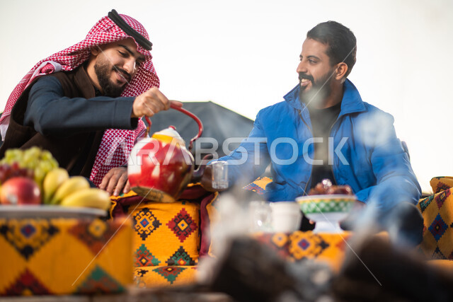 صب الشاي للضيوف، حسن الكرم والضيافة، الجلسات الهادئة في البراري أمام موقد النار، الاستمتاع بالمشروبات الساخنة في الأجواء الشتوية، صورة مقربة لرجلان عربيان خليجيان سعوديان يرتديان الثوب التقليدي والشماغ مع ملابس الشتاء الدافئة يقضون أوقات ممتعة، كشتة بر شبابية