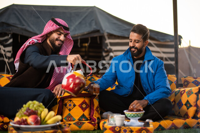 تقديم المشروبات الساخنة في الأجواء الشتوية، كشتة بر شبابية، صب الشاي للضيوف، حسن الكرم والضيافة، الجلسات الهادئة في البراري أمام موقد النار، صورة مقربة لرجلان عربيان خليجيان سعوديان يرتديان الثوب التقليدي والشماغ مع ملابس الشتاء الدافئة يقضون أوقات ممتعة