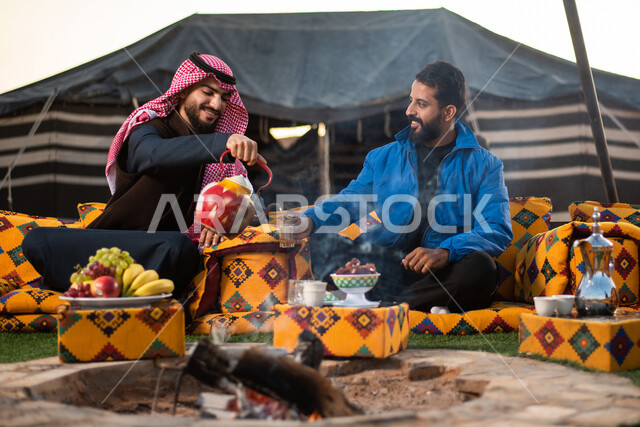 الاستمتاع بالمشروبات الساخنة في الأجواء الشتوية، كشتة بر شبابية، صب الشاي للضيوف، حسن الكرم والضيافة، الجلسات الهادئة في البراري أمام موقد النار، صورة مقربة لرجلان عربيان خليجيان سعوديان يرتديان الثوب التقليدي والشماغ مع ملابس الشتاء الدافئة يقضون أوقات ممتعة