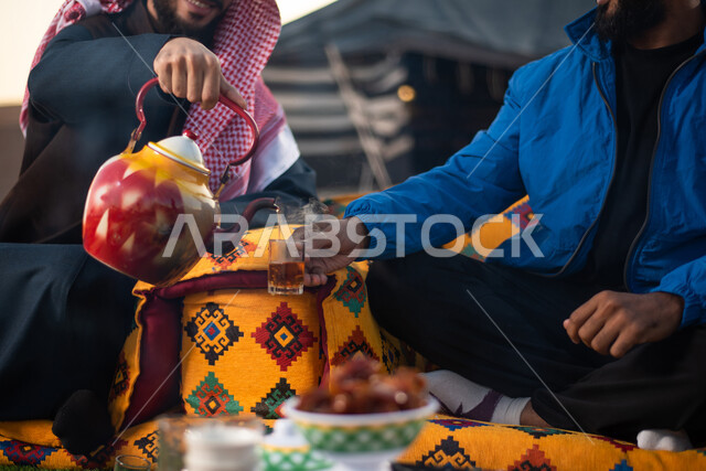 حسن الكرم والضيافة، صب الشاي للضيوف، الجلسات الهادئة في البراري أمام موقد النار، الاستمتاع بالمشروبات الساخنة في الأجواء الشتوية، صورة مقربة لرجلان عربيان خليجيان سعوديان يرتديان الثوب التقليدي والشماغ مع ملابس الشتاء الدافئة يقضون أوقات ممتعة، كشتة بر شبابية
