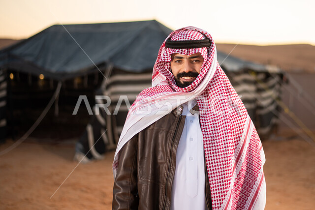 الاهتمام بالمظهر الخارجي، صورة مقربة لرجل عربي خليجي سعودي يرتدي الثوب التقليدي والشماغ مع جاكيت بني جلد ينظر إلى الكاميرا بإيماءات السعادة والسرور، كشتة بر عربية شبابية مسائية، مخيمات سياحية شعبية في صحاري السعودية، اجواء شتوية ليلية