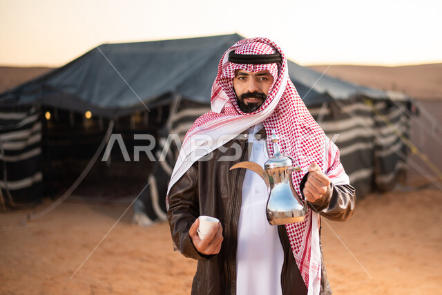 رحلة تخييم برية في الصحراء، صورة مقربة لرجل عربي خليجي سعودي يرتدي الثوب التقليدي مع ملابس الشتاء الدافئة يمسك بيده دلة القهوة العربية، مخيمات سياحية في صحاري السعودية، كشتة بر عربية شبابية، تقديم المشروبات الساخنة للضيوف،حسن الضيافة والاستقبال عند العرب