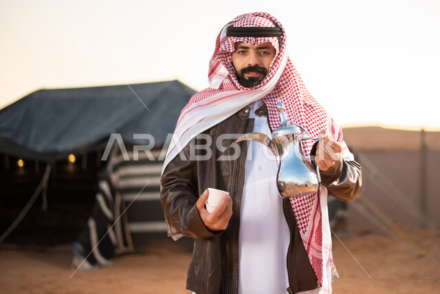 تقديم المشروبات الساخنة للضيوف، حسن الضيافة والاستقبال عند العرب، رحلة تخييم برية في الصحراء، صورة مقربة لرجل عربي خليجي سعودي يرتدي الثوب التقليدي مع ملابس الشتاء الدافئة يمسك بيده دلة القهوة العربية، مخيمات سياحية في صحاري السعودية، كشتة بر عربية شبابية