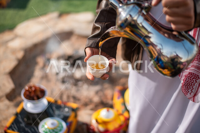 مفهوم إكرام الضيف، صب القهوة من الدلة النحاسية، قضاء أوقات ممتعة مع الأصدقاء، إيماءات البشاشة والترحيب، خيام شعبية في صحاري السعودية، صورة مقربة لرجلان عربيان خليجيان سعوديان يرتديان الثوب التقليدي مع ملابس الشتاء في رحلة تخييم برية، جلسة بر عربية شعبية