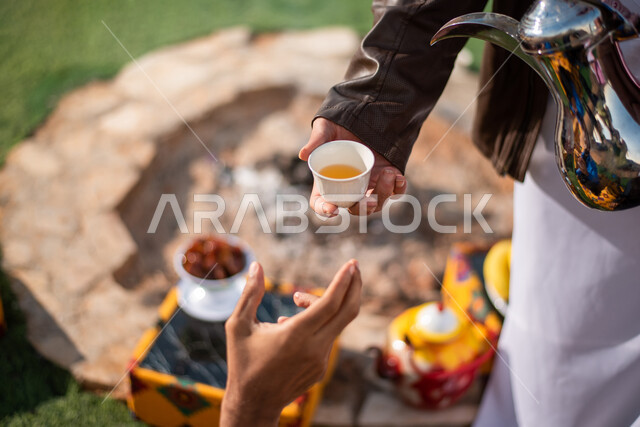 صب القهوة من الدلة النحاسية، قضاء أوقات ممتعة مع الأصدقاء، إيماءات البشاشة والترحيب، خيام شعبية في صحاري السعودية، الكرم والضيافة عند العرب، صورة مقربة لرجلان عربيان خليجيان سعوديان يرتديان الثوب التقليدي مع ملابس الشتاء في رحلة تخييم برية، جلسة بر عربية شعبية