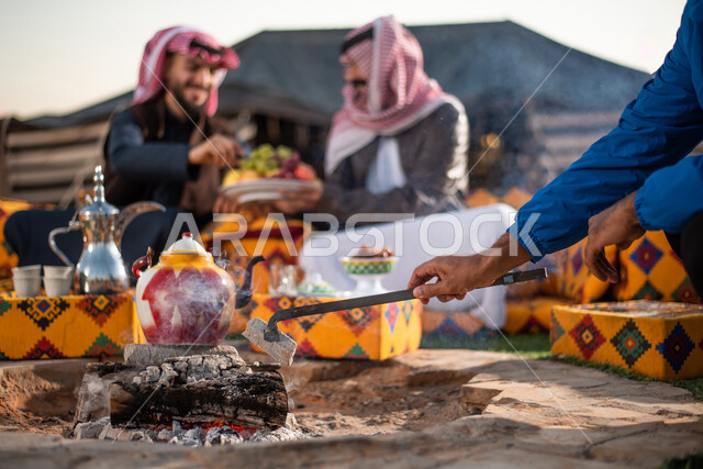 الجلسات الهادئة أمام موقد النار، تقديم الفواكه للضيوف، الكشتات الشعبية في المخيمات في صحاري السعودية، صورة مقربة لرجلان عربيان خليجيان سعوديان يرتديان الثوب التقليدي مع ملابس الشتاء في رحلة تخييم برية، حسن الضيافة والإستقبال، قضاء أوقات ممتعة مع الأصدقاء