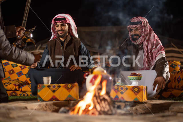 Arab hospitality, serving hot drinks, pouring coffee from a copper pot, a close-up of two Saudi Gulf Arab men wearing traditional thobes and winter clothes on a wild camping trip, a popular Arab camping session, popular tents in the deserts of Saudi Arabia, spending fun times with friends