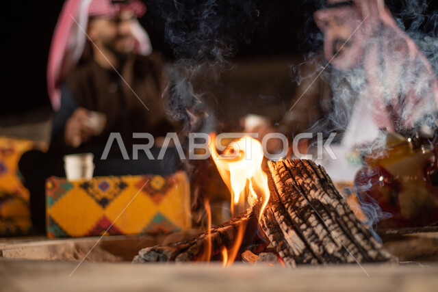 حوارات ونقاشات بين الاصدقاء، الجلسات الهادئة في البراري أمام موقد النار، مفهوم حسن الضيافه والكرم، صورة مقربة لشابان عربيان خليجيان سعوديان يرتديان الثوب التقليدي والشماغ مع ملابس الشتاء في رحلة تخييم برية في صحراء السعودية، تقديم القهوة العربية الساخنة للضيوف