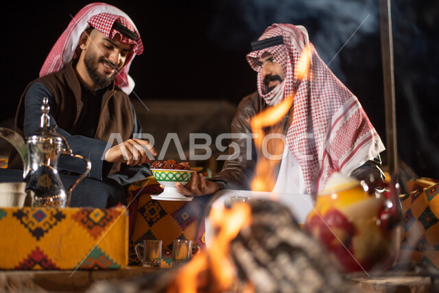 دلة قهوة عربية نحاسية، تقديم التمر للضيوف، مفهوم إكرام الضيوف، رجلان عربيان خليجيان سعوديان يرتديان الثوب التقليدي مع ملابس الشتاء الدافئة يجلسان أمام شغلة النار، وجبة ذات قيمة غذائية عالية، مخيمات سياحية في صحاري السعودية، الاستمتاع في قضاء الوقت مع الأصحاب